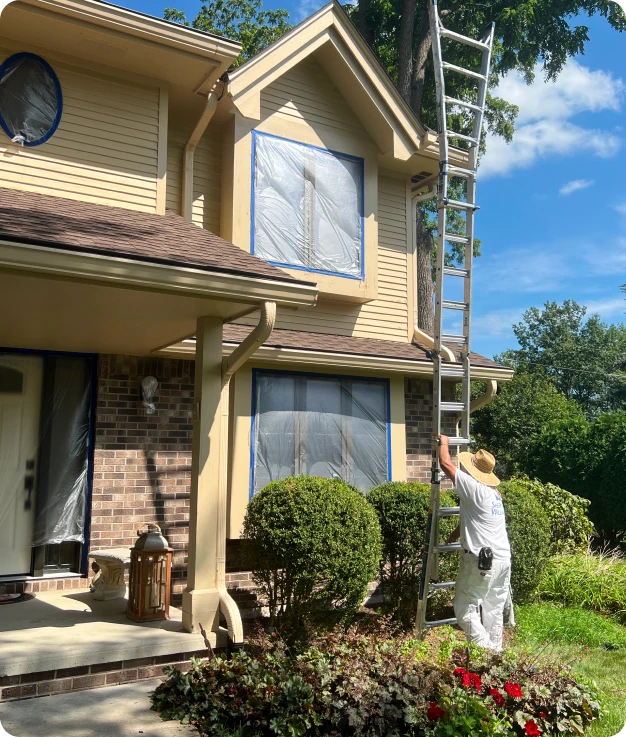 Person in a white uniform setting up a ladder against a house with partially covered windows, preparing for a painting project, surrounded by well-maintained landscaping in a residential area.