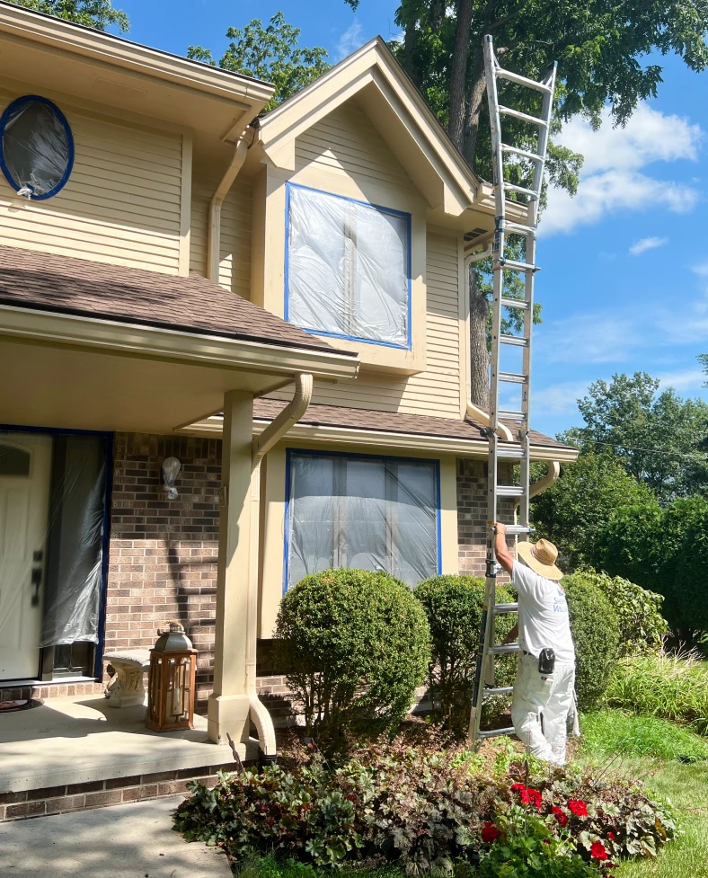Man in white painting attire setting up a ladder against a house with covered windows, preparing for exterior painting, surrounded by well-maintained landscaping in Rockford, IL.